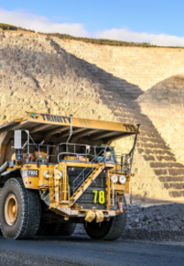 A large mining truck labeled 'Trinity' driving near a steep rocky slope under a blue sky.